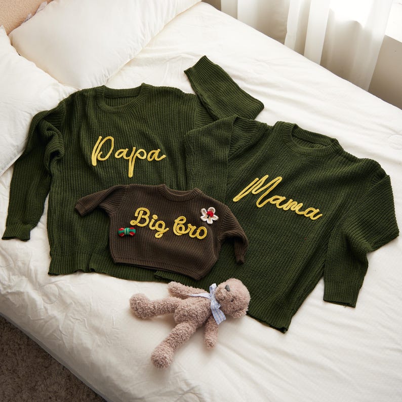 May include: Green knit sweaters with yellow embroidered text reading "Papa" and "Mama". A smaller brown sweater says "Big bro". A teddy bear lies nearby. The sweaters are on a white bed.