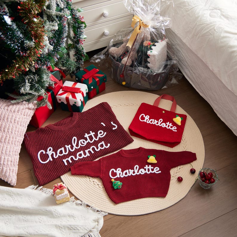 May include: A festive scene with Christmas gifts and personalized items. A burgundy sweater and a matching tote bag are embroidered with the name "Charlotte". A smaller sweater is also personalized. A gift basket and a Christmas tree are in the background.