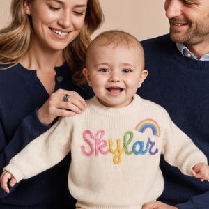 Op de afbeelding: Een cr&egrave;mekleurige gebreide trui met lange mouwen en de naam Skylar geborduurd in gekleurd garen op de borst, vergezeld van een kleine regenboogafbeelding boven de tekst. De trui heeft een geribbelde ronde hals, boorden en een zoom.