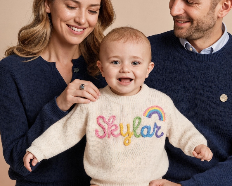 Op de afbeelding: Een cr&egrave;mekleurige gebreide trui met lange mouwen en de naam Skylar geborduurd in gekleurd garen op de borst, vergezeld van een kleine regenboogafbeelding boven de tekst. De trui heeft een geribbelde ronde hals, boorden en een zoom.