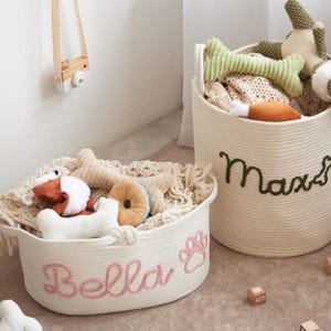 Puede incluir: Dos cestas de cuerda color crema llenas de juguetes para perros. Una cesta tiene "Bella" bordado en rosa, con una huella de pata. La otra tiene "Max" bordado en verde. Los juguetes incluyen huesos, una rosquilla y peluches.
