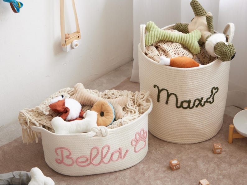 Puede incluir: Dos cestas de cuerda color crema llenas de juguetes para perros. Una cesta tiene "Bella" bordado en rosa, con una huella de pata. La otra tiene "Max" bordado en verde. Los juguetes incluyen huesos, una rosquilla y peluches.