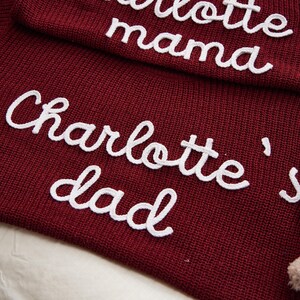 May include: Close-up of a burgundy knit fabric with white embroidered text. The text reads "Charlotte's dad" and "mama". The fabric has a textured appearance, and the embroidery is in a cursive style.