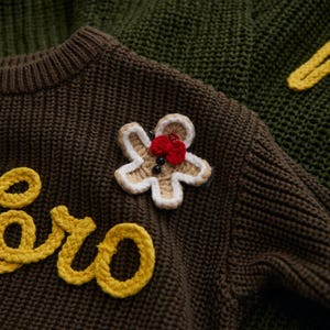 May include: Close-up of a brown knit sweater with a gingerbread man applique. The gingerbread man is beige with white trim, a red bow, and black button eyes. Yellow text is visible on the sweater.