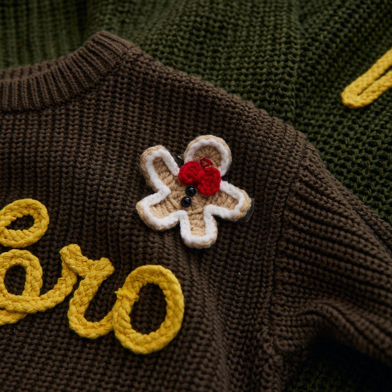 May include: Close-up of a brown knit sweater with a gingerbread man applique. The gingerbread man is beige with white trim, a red bow, and black button eyes. Yellow text is visible on the sweater.