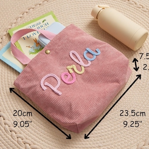 Może przedstawiać: R&oacute;żowa torba typu tote z sztruksu z haftowanym imieniem "Perla" w kolorowych literach. Torba o wymiarach 23,5 cm x 20 cm x 7,5 cm zawiera książki i jasnoniebieskie etui. Obok torby leży beżowa butelka na wodę. Torba ma r&oacute;żowy uchwyt i złoty guzik.