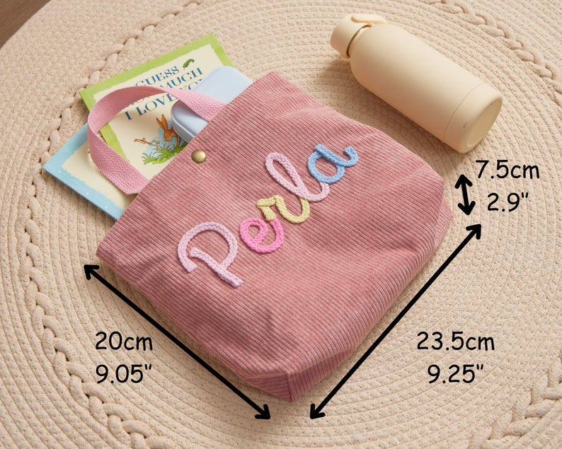 Może przedstawiać: R&oacute;żowa torba typu tote z sztruksu z haftowanym imieniem "Perla" w kolorowych literach. Torba o wymiarach 23,5 cm x 20 cm x 7,5 cm zawiera książki i jasnoniebieskie etui. Obok torby leży beżowa butelka na wodę. Torba ma r&oacute;żowy uchwyt i złoty guzik.