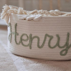 Puede incluir: Una cesta de almacenamiento blanca tejida con el nombre "Henry" bordado en verde en cursiva. La cesta est&aacute; llena de textiles blancos suaves y tiene una borla decorativa en el lateral. La cesta est&aacute; sobre una alfombra de color claro.