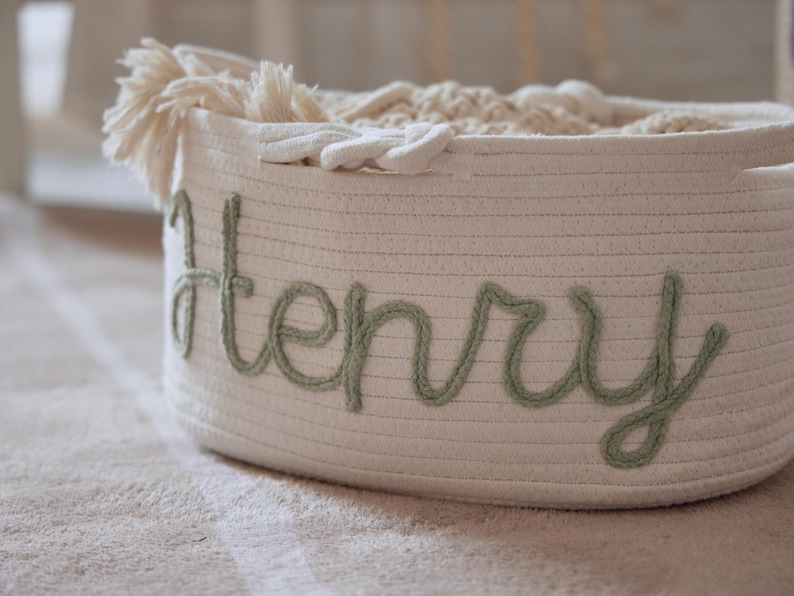 Puede incluir: Una cesta de almacenamiento blanca tejida con el nombre "Henry" bordado en verde en cursiva. La cesta est&aacute; llena de textiles blancos suaves y tiene una borla decorativa en el lateral. La cesta est&aacute; sobre una alfombra de color claro.