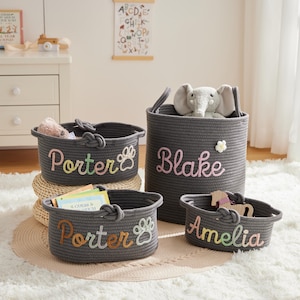 Peut inclure: Quatre paniers de rangement en corde grise de différentes tailles. Chaque panier porte un nom brodé en fil coloré. Les noms sont Porter, Blake et Amelia. Un panier a une empreinte de patte. Les paniers sont sur un tapis blanc.