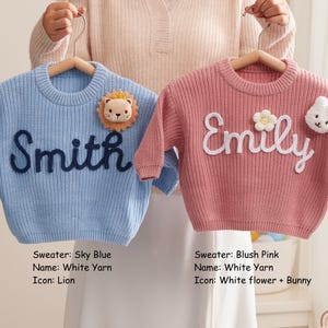 Op de afbeelding: Twee gebreide truien met lange mouwen op houten kleerhangers. De hemelsblauwe trui heeft de naam Smith in donkerblauw garen en een leeuw-icoon. De oudroze trui heeft de naam Emily in wit garen met een witte bloem en een wit konijn-icoon.
