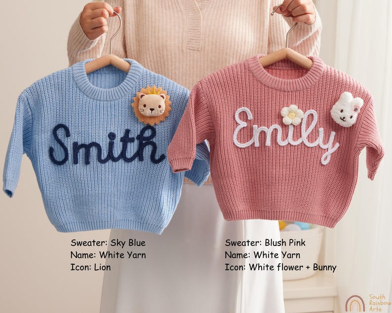Op de afbeelding: Twee gebreide truien met lange mouwen op houten kleerhangers. De hemelsblauwe trui heeft de naam Smith in donkerblauw garen en een leeuw-icoon. De oudroze trui heeft de naam Emily in wit garen met een witte bloem en een wit konijn-icoon.
