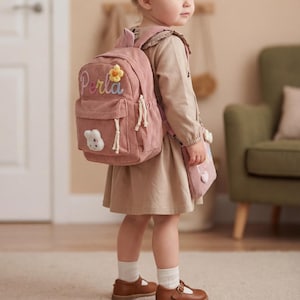 Peut inclure: Un sac à dos en velours côtelé rose avec le nom "Perla" brodé en cursive, une fleur et une application de lapin. Le sac à dos a des cordons blancs et une petite pochette assortie. L'enfant porte des chaussures marron et des chaussettes blanches.