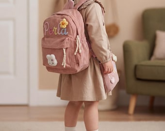 May include: A pink corduroy backpack with the name "Perla" embroidered in cursive, a flower, and a bunny applique. The backpack has white drawstrings and a matching small purse. The child is wearing brown shoes and white socks.