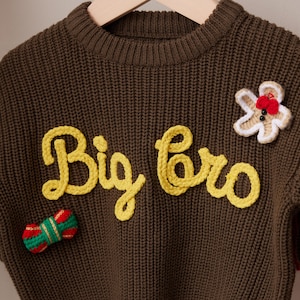May include: A brown knit sweater with the words "Big Bro" in yellow script. The sweater has a gingerbread man and a wrapped candy applique. The sweater is hanging on a wooden hanger.