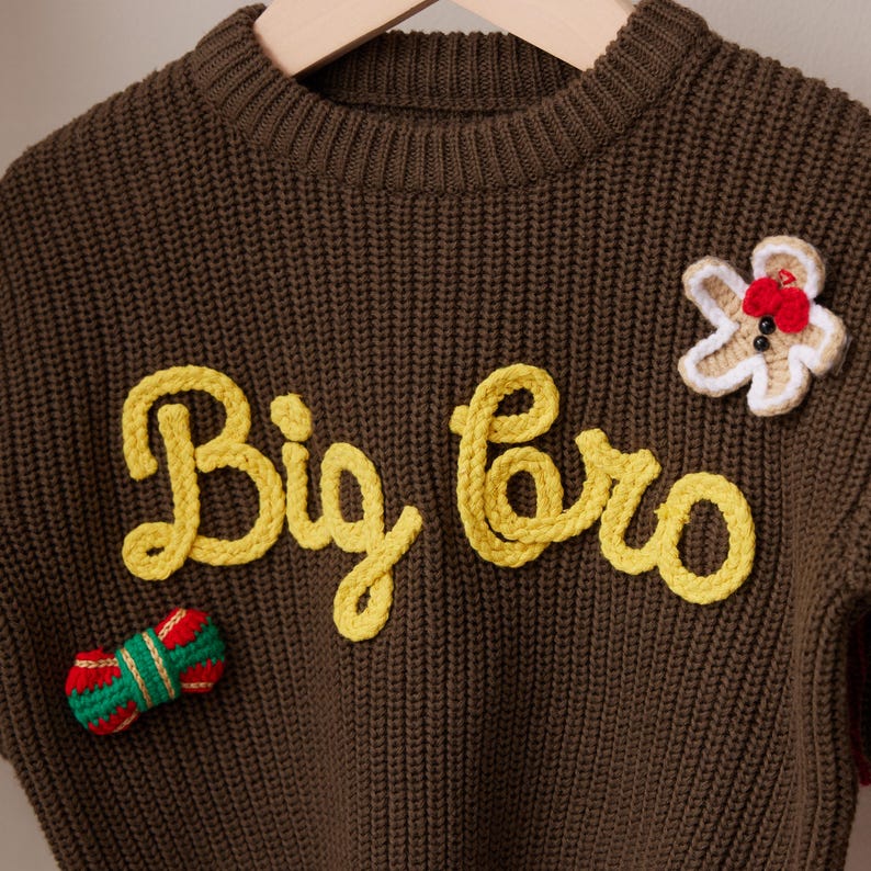 May include: A brown knit sweater with the words "Big Bro" in yellow script. The sweater has a gingerbread man and a wrapped candy applique. The sweater is hanging on a wooden hanger.