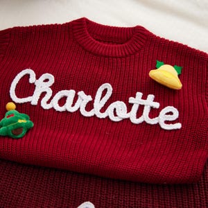 May include: A red knit sweater with the name "Charlotte" in white script across the front. The sweater features a small green Christmas tree and a yellow bell applique. The sweater is folded.