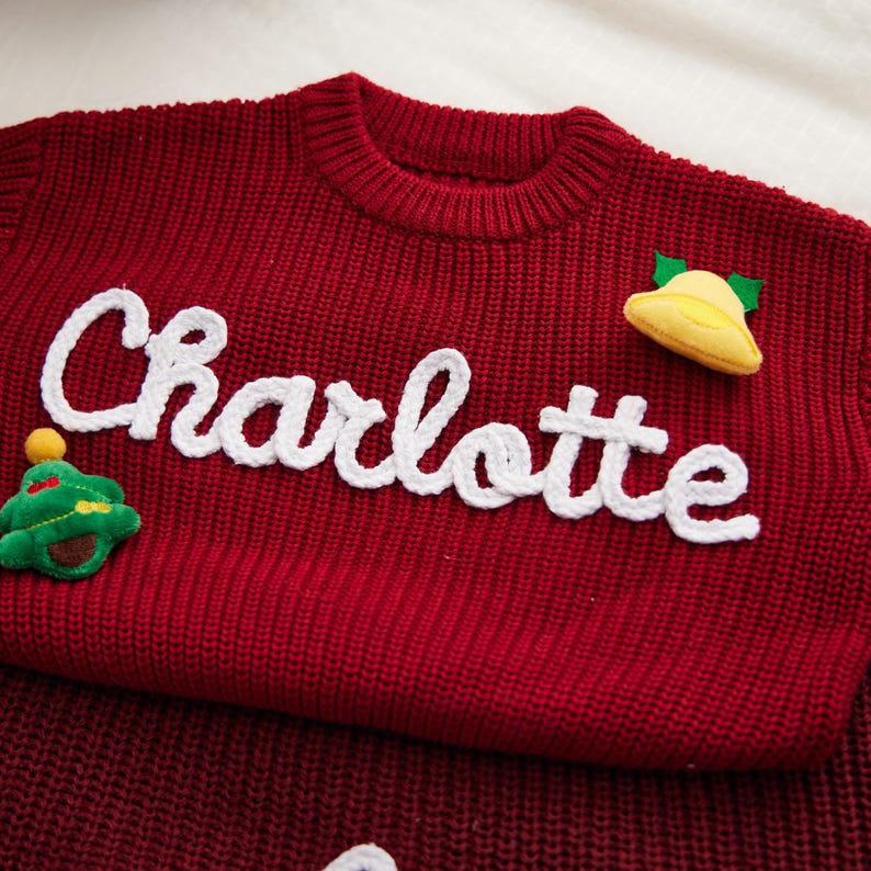May include: A red knit sweater with the name "Charlotte" in white script across the front. The sweater features a small green Christmas tree and a yellow bell applique. The sweater is folded.