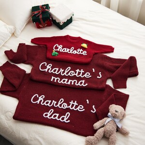 May include: A family matching sweater set in burgundy red. The set includes sweaters with the names "Charlotte", "Charlotte's mama", and "Charlotte's dad" in white script. A small teddy bear and wrapped gifts are also in the image.