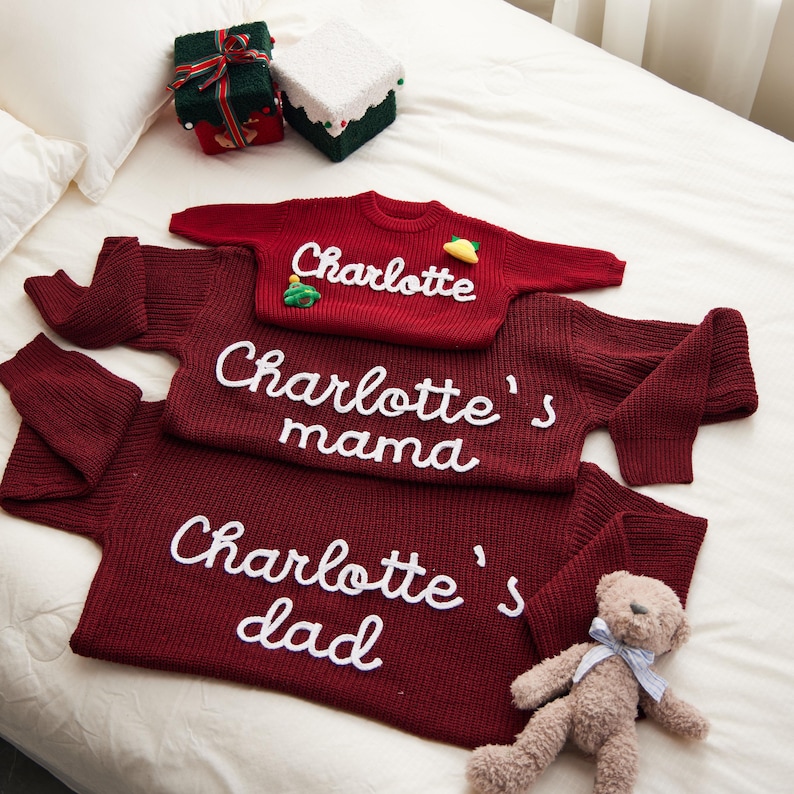 May include: A family matching sweater set in burgundy red. The set includes sweaters with the names "Charlotte", "Charlotte's mama", and "Charlotte's dad" in white script. A small teddy bear and wrapped gifts are also in the image.