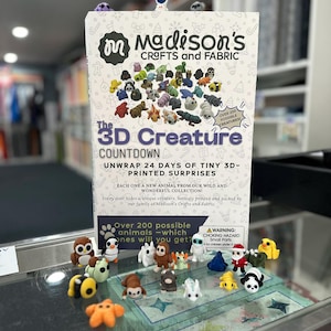 May include: A display featuring a box labeled "Madison's Crafts and Fabric" with "The 3D Creature Countdown" text. The box is surrounded by small, colorful 3D-printed animal figurines, including an owl, penguin, and panda.