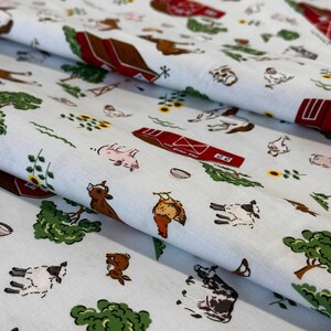 Könnte beinhalten: Weißer Stoff mit einem Bauernhofmuster. Das Design umfasst rote Scheunen, Nutztiere wie Kühe, Schweine und Schafe sowie Sonnenblumen. Der Stoff ist mit braunen, roten, grünen, gelben und rosa Elementen gemustert.