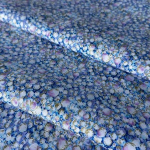 以下が含まれることがあります： 白、青、紫の円が金色のアクセントで繰り返されるパターンの生地のクローズアップ。生地は折りたたまれ、デザインの質感と深さを示しています。全体的なカラーパレットはクールで落ち着いています。