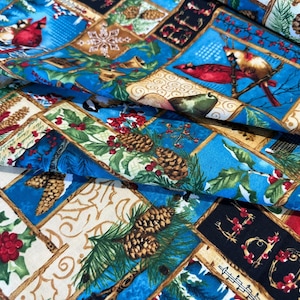 May include: Close-up of a fabric with a patchwork design featuring winter and Christmas motifs. The fabric has a blue background with squares of red cardinals, pinecones, holly, and the word "BELIEVE". The fabric is folded.