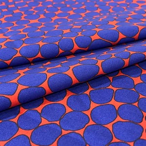 May include: A fabric with a repeating pattern of blue, rounded shapes on a red background. The fabric is folded, showing the texture and drape of the material. The design is reminiscent of pebbles or organic forms.