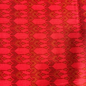 May include: A close-up of red fabric featuring a repeating pattern of stylised floral and diamond shapes in brown and rust hues. The fabric has a textured appearance, possibly woven. The design is symmetrical and visually engaging.