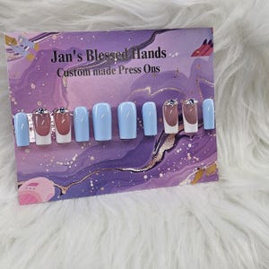 May include: A set of custom-made press-on nails. The nails are light blue, with some featuring a white and brown French tip design. Some nails have rhinestones. The card reads "Jan's Blessed Hands Custom made Press Ons."