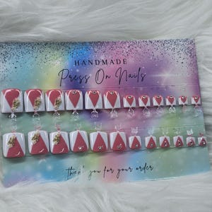 May include: A set of 10 pink and white press-on nails with gold accents and rhinestones. The nails are arranged on a card that says "Handmade Press On Nails" and "Thank you for your order".