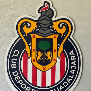 May include: A layered wooden sign featuring the Club Deportivo Guadalajara crest. The crest includes a shield with a tree and lions, surrounded by a navy blue ring with white text. The name "VELAZQUEZ" is displayed below in red letters.