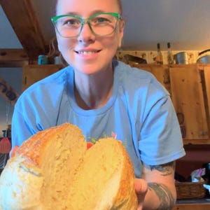 Könnte beinhalten: Ein frisch gebackenes Brot, halbiert, wodurch ein weiches, luftiges Inneres sichtbar wird. Das Brot hat eine goldbraune Kruste. Die Person, die das Brot hält, trägt ein blaues T-Shirt und eine grüne Brille.