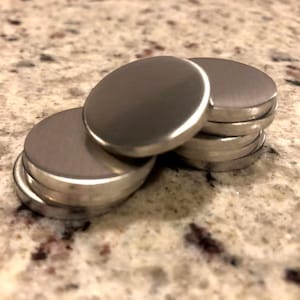 May include: Several small, round, silver-coloured metal objects are stacked. The objects are on top of each other, with some slightly askew. The surface has a brushed metal appearance, and the edges are smooth and rounded.
