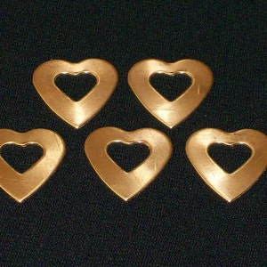 May include: Five heart-shaped metal charms with a cutout heart in the center. The charms are a warm copper color and are arranged on a dark background. These charms could be used for jewelry making or other craft projects.