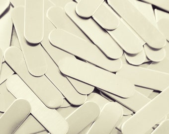May include: A close-up shot of a pile of light-colored, oval-ended wooden craft sticks. The sticks are scattered and overlapping, creating a textured surface. The color is a muted, off-white, with subtle variations in tone.
