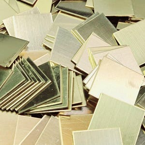 May include: A pile of small, square brass metal sheets. The sheets have a brushed finish and vary in colour, from gold to a muted brass tone. Some are stacked neatly, while others are scattered.