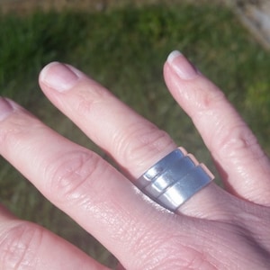 May include: A silver-colored ring with a stepped design is worn on a finger. The ring has a modern, geometric style, reflecting light. The ring's design features three stacked, rectangular bands, creating a unique and eye-catching accessory.