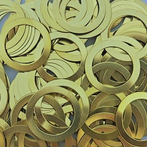 May include: A collection of brass-colored, circular washers. The washers are flat with a rounded outer edge and a circular opening in the center. The washers are scattered and overlapping, creating a textured surface.