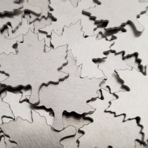 May include: A collection of silver-colored metal maple leaf cutouts. The leaves have a detailed, textured surface and a jagged edge. The leaves are scattered across a surface, creating a pattern.