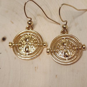 May include: Gold-tone earrings with a circular design featuring a textured center and concentric circles. The earrings have a hook closure.