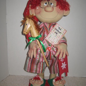 May include: A fabric doll with red hair, glasses, and a red nightcap, wearing striped pajamas in red, green, and white. The doll holds a wooden hobby horse and a book titled The Littlest Elf, while wearing elf-faced slippers on a red base.