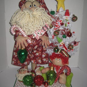 May include: A fabric Santa Claus doll with a yarn beard, glasses, and a red Santa-print outfit, holding a candy cane and a green lightbulb ornament. Beside it stands a white Christmas tree with ornaments, a yellow star, and a small elf doll.