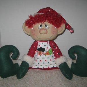May include: A fabric elf doll with a red hat, red hair, and a tan face. The doll wears a red long-sleeved shirt with white faux fur cuffs, dark green mittens, and dark green curled shoes. A white apron with red and green polka dots features a pocket.