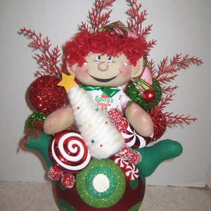 May include: A fabric elf doll with red hair and a white shirt printed with the text Santa's Sidekick. The doll holds a white cone-shaped tree with a yellow star and is surrounded by red and green glittery ornaments, red sprigs, and candy-themed decorations.