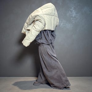 Op de afbeelding: Een crèmekleurige, gewatteerde pufferjas wordt gedragen over een grijze hoodie en een broek met wijde pijpen. De broek en hoodie hebben een vergelijkbare grijstint. De outfit wordt gecombineerd met grijze laarzen. De achtergrond is effen grijs.