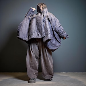 May include: A person wearing a dark blue, oversized quilted jacket with a high collar and wide, matching trousers. The jacket has a unique, cape-like design. The trousers are a muted grey colour. The outfit is a modern take on outerwear.