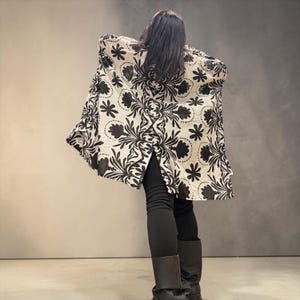 Könnte beinhalten: Eine langärmelige, vorne offene Jacke mit cremefarbener Basis und einem auffälligen, sich wiederholenden schwarzen Blumen- und Damastmuster. Das Kleidungsstück hat eine lockere, fließende Silhouette, einen geraden Saum und wird über einer schwarzen Leggings und dunklen Stiefeln getragen.