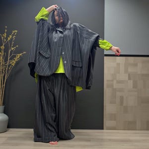以下が含まれることがあります： グレーのピンストライプのスーツ。大きなポケットが付いたケープのようなジャケットとワイドレッグパンツで構成されています。明るい緑色の長袖シャツの上に着用されています。この服装はモダンでアバンギャルドなファッションです。